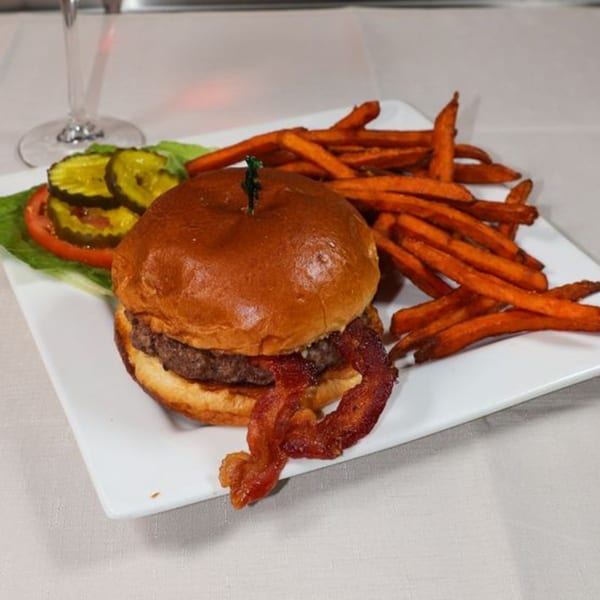 A Prime 7 bacon cheeseburger with sweet potato fries, pickles, and tomato on a white plate.