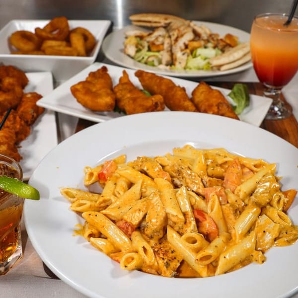 A table laden with American dishes including penne pasta, fried fish, onion rings, salad, and cocktails.
